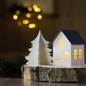 Pode incluir: Uma &aacute;rvore de Natal de papel branco e uma casa de papel branco com telhado azul ficam em cima de uma fatia de madeira. A casa tem janelas e uma porta com uma luz brilhando atrav&eacute;s.
