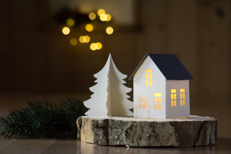 Pode incluir: Uma &aacute;rvore de Natal de papel branco e uma casa de papel branco com telhado azul ficam em cima de uma fatia de madeira. A casa tem janelas e uma porta com uma luz brilhando atrav&eacute;s.