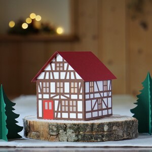 May include: A papercraft model of a small, white and brown house with a red roof. The house is sitting on a wooden log slice with two green papercraft trees on either side.