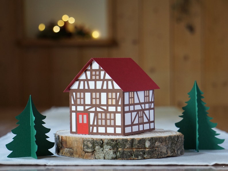 May include: A papercraft model of a small, white and brown house with a red roof. The house is sitting on a wooden log slice with two green papercraft trees on either side.