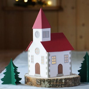 May include: A white paper church with a red roof and a clock on the front. The church is sitting on a wooden log with two green paper trees on either side.