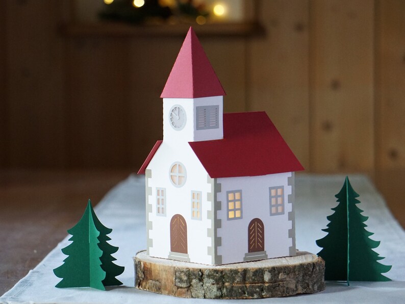 May include: A white paper church with a red roof and a clock on the front. The church is sitting on a wooden log with two green paper trees on either side.