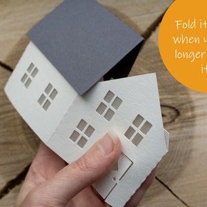 Pode incluir: Uma casa de papel branco com um telhado de papel cinza. A casa tem janelas recortadas e est&aacute; parcialmente dobrada. O texto "Dobre-a na horizontal quando n&atilde;o precisar mais dela" est&aacute; escrito em um c&iacute;rculo amarelo.