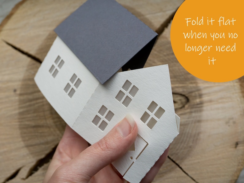 Pode incluir: Uma casa de papel branco com um telhado de papel cinza. A casa tem janelas recortadas e est&aacute; parcialmente dobrada. O texto "Dobre-a na horizontal quando n&atilde;o precisar mais dela" est&aacute; escrito em um c&iacute;rculo amarelo.