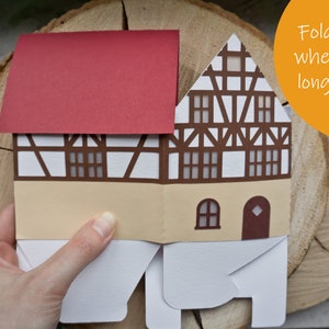 May include: A papercraft house with a red roof, white walls, and brown trim. The house is folded flat and has a text label that says "Fold it flat when you no longer need it".