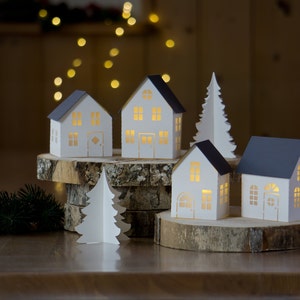 Pode incluir: Um conjunto de cinco casas de papel branco com janelas e portas recortadas, iluminadas por dentro, dispostas em uma base de madeira com uma pequena &aacute;rvore de Natal de papel na frente.