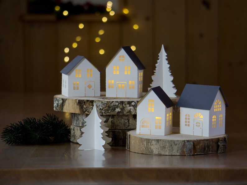 Pode incluir: Um conjunto de cinco casas de papel branco com janelas e portas recortadas, iluminadas por dentro, dispostas em uma base de madeira com uma pequena &aacute;rvore de Natal de papel na frente.