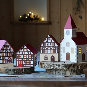 May include: A set of four papercraft houses, including a church with a clock tower, on wooden bases. The houses are decorated with white, brown, and red accents. Two small green paper trees are on either side of the houses.