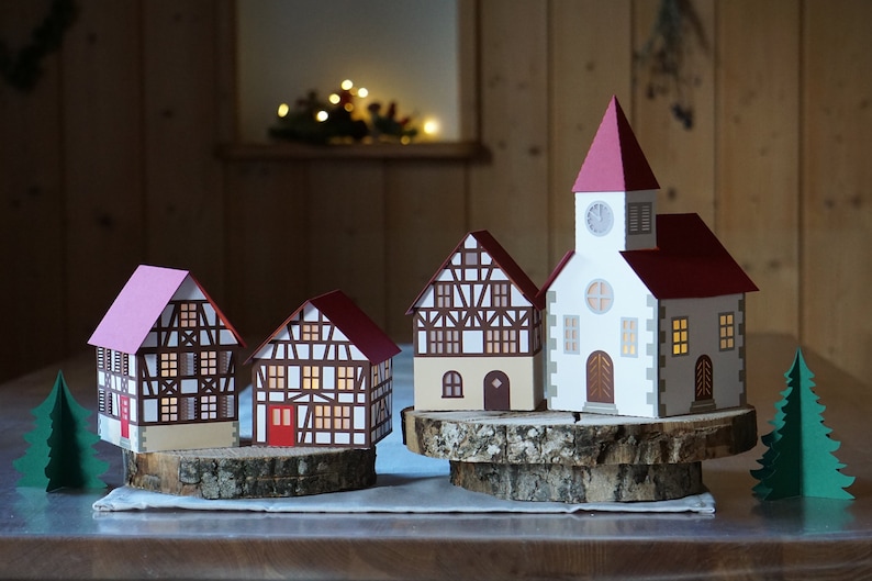 May include: A set of four papercraft houses, including a church with a clock tower, on wooden bases. The houses are decorated with white, brown, and red accents. Two small green paper trees are on either side of the houses.