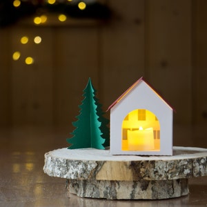 May include: A white paper house with a red roof and a lit candle inside, next to a green paper Christmas tree on a wooden slice.