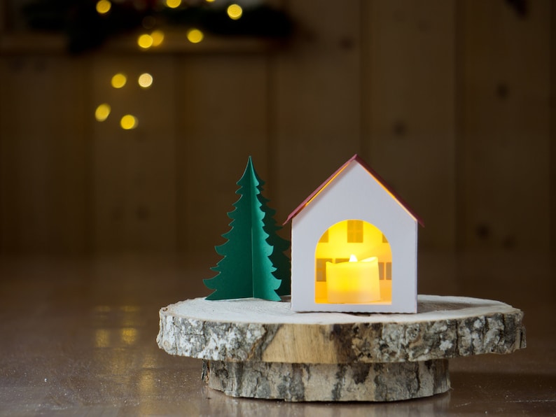 May include: A white paper house with a red roof and a lit candle inside, next to a green paper Christmas tree on a wooden slice.