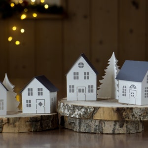 Pode incluir: Cinco casas de papel e uma &aacute;rvore de papel, todas brancas com detalhes cinzentos, est&atilde;o dispostas em tr&ecirc;s fatias de madeira. As casas t&ecirc;m janelas e portas, e a &aacute;rvore tem um topo pontudo.
