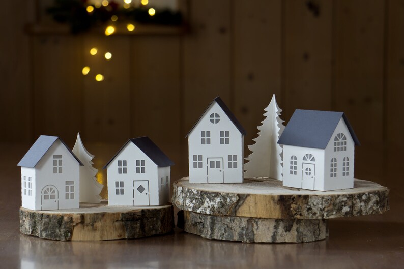 Pode incluir: Cinco casas de papel e uma &aacute;rvore de papel, todas brancas com detalhes cinzentos, est&atilde;o dispostas em tr&ecirc;s fatias de madeira. As casas t&ecirc;m janelas e portas, e a &aacute;rvore tem um topo pontudo.