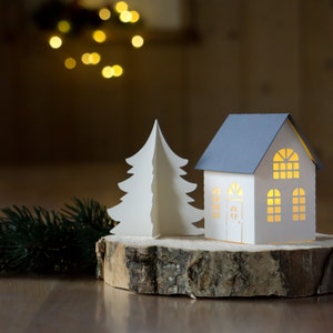 Pode incluir: Uma &aacute;rvore de Natal de papel branco e uma casa de papel branco com telhado cinza est&atilde;o sobre uma fatia de madeira. A casa tem janelas com um brilho quente vindo do interior.