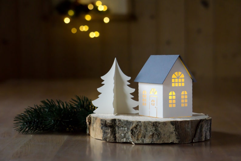 Pode incluir: Uma &aacute;rvore de Natal de papel branco e uma casa de papel branco com telhado cinza est&atilde;o sobre uma fatia de madeira. A casa tem janelas com um brilho quente vindo do interior.