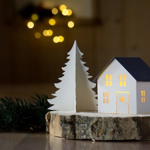 Pode incluir: Uma &aacute;rvore de Natal de papel branco e uma casa de papel branco com telhado azul, ambas iluminadas por dentro, s&atilde;o exibidas em uma fatia de madeira. Um ramo de pinheiro verde est&aacute; em primeiro plano.