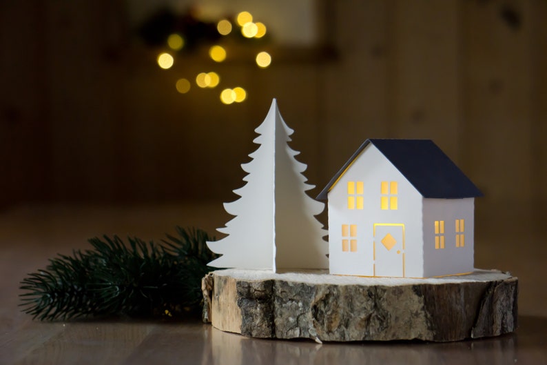 Pode incluir: Uma &aacute;rvore de Natal de papel branco e uma casa de papel branco com telhado azul, ambas iluminadas por dentro, s&atilde;o exibidas em uma fatia de madeira. Um ramo de pinheiro verde est&aacute; em primeiro plano.