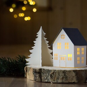 Pode incluir: Uma casa de papel branco com telhado azul e uma janela iluminada est&aacute; sobre uma fatia de madeira ao lado de uma &aacute;rvore de Natal de papel branco.