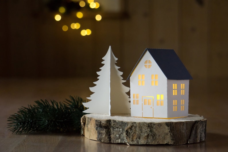 Pode incluir: Uma casa de papel branco com telhado azul e uma janela iluminada est&aacute; sobre uma fatia de madeira ao lado de uma &aacute;rvore de Natal de papel branco.