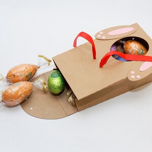 Puede incluir: Una bolsa de papel marr&oacute;n con orejas de conejo y asas de cinta roja. La bolsa est&aacute; abierta y llena de huevos de Pascua de chocolate, incluyendo uno verde con papel de aluminio dorado.