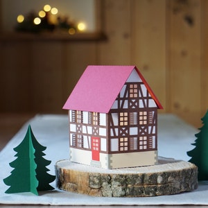 May include: A papercraft model of a traditional German half-timbered house with a red roof, white walls, and brown beams. The house is sitting on a wooden slice with two green papercraft trees on either side.