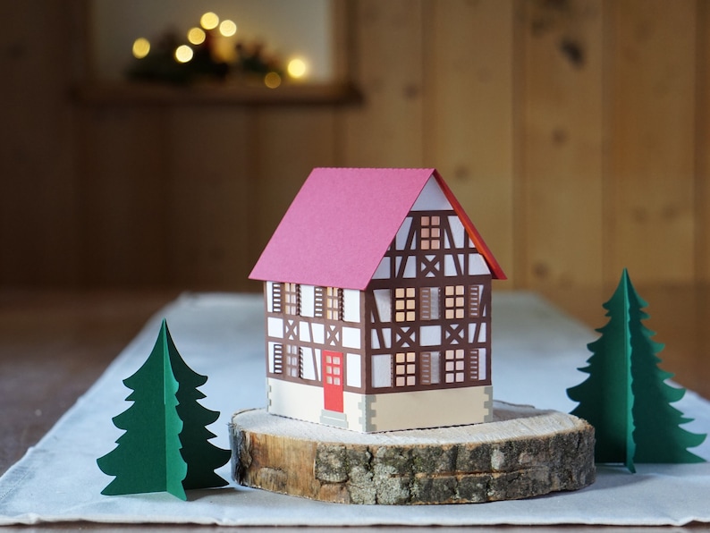 May include: A papercraft model of a traditional German half-timbered house with a red roof, white walls, and brown beams. The house is sitting on a wooden slice with two green papercraft trees on either side.