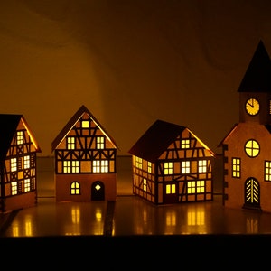 May include: Four illuminated paper houses with a clock tower, all with a traditional German style, with black trim and windows, and a warm yellow glow.