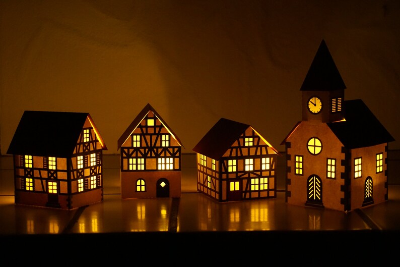 May include: Four illuminated paper houses with a clock tower, all with a traditional German style, with black trim and windows, and a warm yellow glow.
