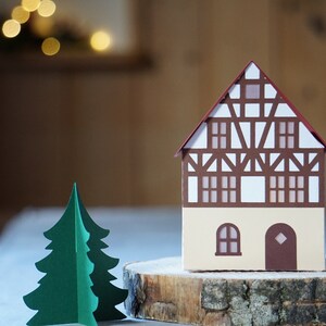 May include: A papercraft model of a brown and white half-timbered house with a red roof, sitting on a wooden log slice. A green papercraft Christmas tree is in the foreground.