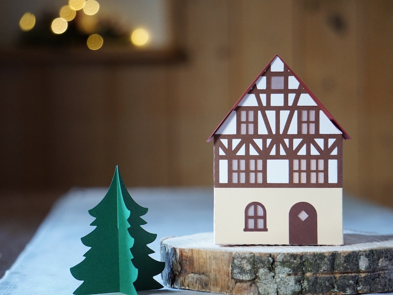 May include: A papercraft model of a brown and white half-timbered house with a red roof, sitting on a wooden log slice. A green papercraft Christmas tree is in the foreground.