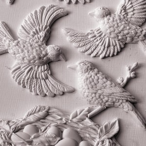 May include: A detailed, monochromatic relief sculpture featuring birds in flight and a nest with eggs. The birds have textured feathers and are depicted in various poses. The nest is intricately woven with leaves and feathers.