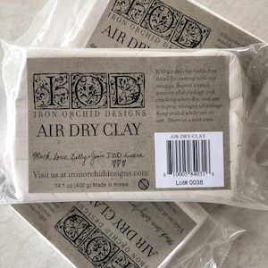 May include: A package of Iron Orchid Designs air dry clay, 14.1 oz (400 g), made in Korea. The package is white with black text and a barcode. The text on the package reads "IOD air dry clay holds fine detail for casting with our moulds. Expect a small amount of shrinkage and cracking when dry, and use it to your vintagey advantage. Keep sealed while not in use. Store in a cool area."