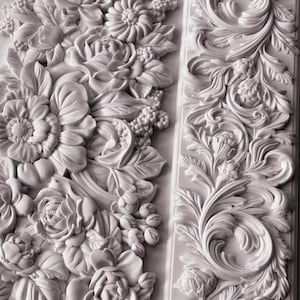 May include: A white architectural molding panel featuring a high-relief carved design. The left section displays a dense arrangement of blooming flowers and leaves, while the right section shows a linear pattern of scrolling acanthus leaves.