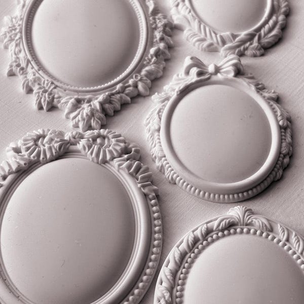 May include: Five white oval and circular frames with raised relief details. Each frame features a smooth center and decorative borders including beaded edges, floral motifs, and a bow accent on one piece. The items are solid white in color.