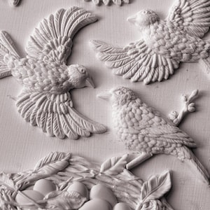May include: A white relief wall art piece featuring three sculpted birds in flight and perched positions, alongside a detailed bird nest containing five smooth, oval-shaped eggs. The textured design highlights individual feathers and twigs.