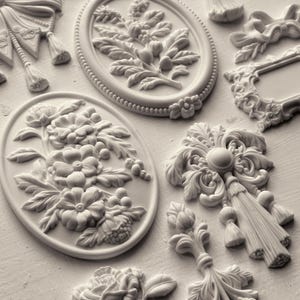 May include: A collection of off-white decorative resin appliques featuring various relief designs including oval floral plaques, bows, tassels, and ornate scrollwork. These individual molded pieces display intricate botanical and architectural details.