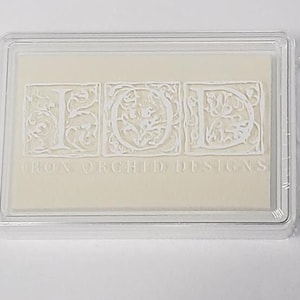 May include: A rectangular clear plastic storage case containing a cream-colored ink pad. The lid features the white embossed letters IOD and the text IRON ORCHID DESIGNS, with each letter enclosed in a square frame with intricate floral scrollwork.