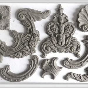 May include: A collection of gray, ornate decorative molding pieces in various shapes, including floral and scroll designs. The pieces are arranged on a white surface, showcasing their intricate details and textures. Ideal for craft projects.