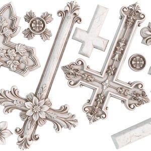 May include: A collection of white crosses and decorative floral elements. The crosses feature intricate designs, including floral and leaf patterns. Some crosses have a distressed, aged appearance, while others are more ornate.