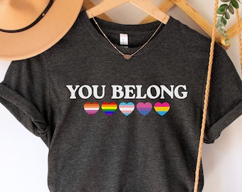 Puede incluir: Camiseta de color gris oscuro jaspeado con el texto "YOU BELONG" y cinco corazones arcoíris. La camiseta está en una percha con un sombrero de paja y una bolsa tejida.