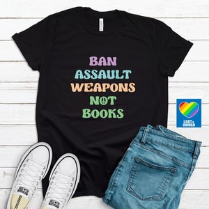 May include: Black t-shirt with the text "BAN ASSAULT WEAPONS NOT BOOKS" in pastel colors. The shirt is paired with blue jeans and white sneakers. A rainbow heart logo with "LGBT+ OWNED" is on the right.