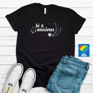 Pode incluir: Camiseta preta com o texto branco "bi & anxious", acompanhada de gr&aacute;ficos de estrelas e tra&ccedil;os. A camiseta &eacute; combinada com jeans azuis e t&ecirc;nis brancos. Um logotipo de cora&ccedil;&atilde;o arco-&iacute;ris com "LGBT+ OWNED" &eacute; vis&iacute;vel.