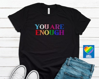 Puede incluir: Camiseta negra con el texto "YOU ARE ENOUGH" en colores arcoíris. La camiseta está colocada sobre una superficie de madera blanca con un par de zapatillas blancas, unos vaqueros azules y un cuadrado azul con un corazón arcoíris y el texto "LGBT+ OWNED".