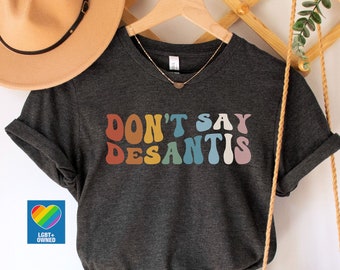 May include: Dark grey heather v-neck t-shirt with the text "DON'T SAY DESANTIS" in retro rainbow colors. The shirt is paired with denim shorts, a tan hat, and a macrame bag. A small rainbow heart logo is visible.