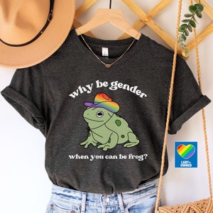 May include: Dark grey heather t-shirt featuring a frog in a rainbow hat with the text "why be gender when you can be frog?" The outfit includes denim shorts, a tan hat, and a macrame bag with a fringed design.