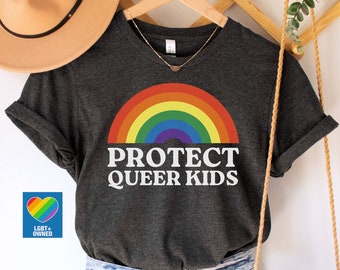 Puede incluir: Camiseta gris oscuro jaspeado con un diseño de arcoíris y las palabras "PROTECT QUEER KIDS" en blanco. La camiseta se combina con pantalones cortos de mezclilla, un sombrero beige y un bolso de macramé. Un logo LGBT+ owned es visible.