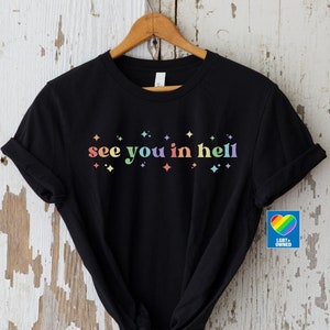 Puede incluir: Camiseta negra con el texto "see you in hell" en colores arco&iacute;ris y peque&ntilde;as estrellas arco&iacute;ris. La camiseta est&aacute; sobre un fondo de madera blanca. Un peque&ntilde;o cuadrado azul con un coraz&oacute;n arco&iacute;ris y el texto "LGBT+ OWNED" est&aacute; en la esquina inferior derecha.