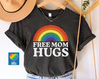 May include: Dark grey heather t-shirt with a rainbow and the text "Free Mom Hugs". The shirt is on a hanger with a straw hat and a woven bag.