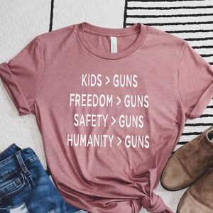 May include: Heather mauve t-shirt with the text "KIDS > GUNS, FREEDOM > GUNS, SAFETY > GUNS, HUMANITY > GUNS" in white. The shirt is styled with denim shorts and brown suede ankle boots on a black and white striped rug.