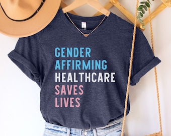 Könnte beinhalten: Heather-Marineblaues T-Shirt mit dem Text "Gender Affirming Healthcare Saves Lives" in hellblau und rosa Schrift. Das Shirt hängt an einem Holzbügel mit einem braunen Hut und einer gewebten Tasche.